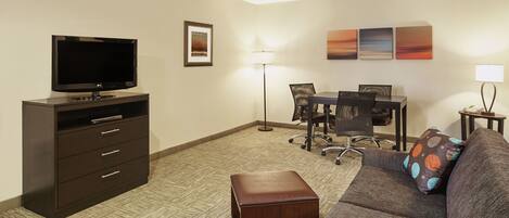 A hotel room with a flat-screen TV, a desk with a chair, a sofa with a pillow, a lamp, and artwork on the wall.