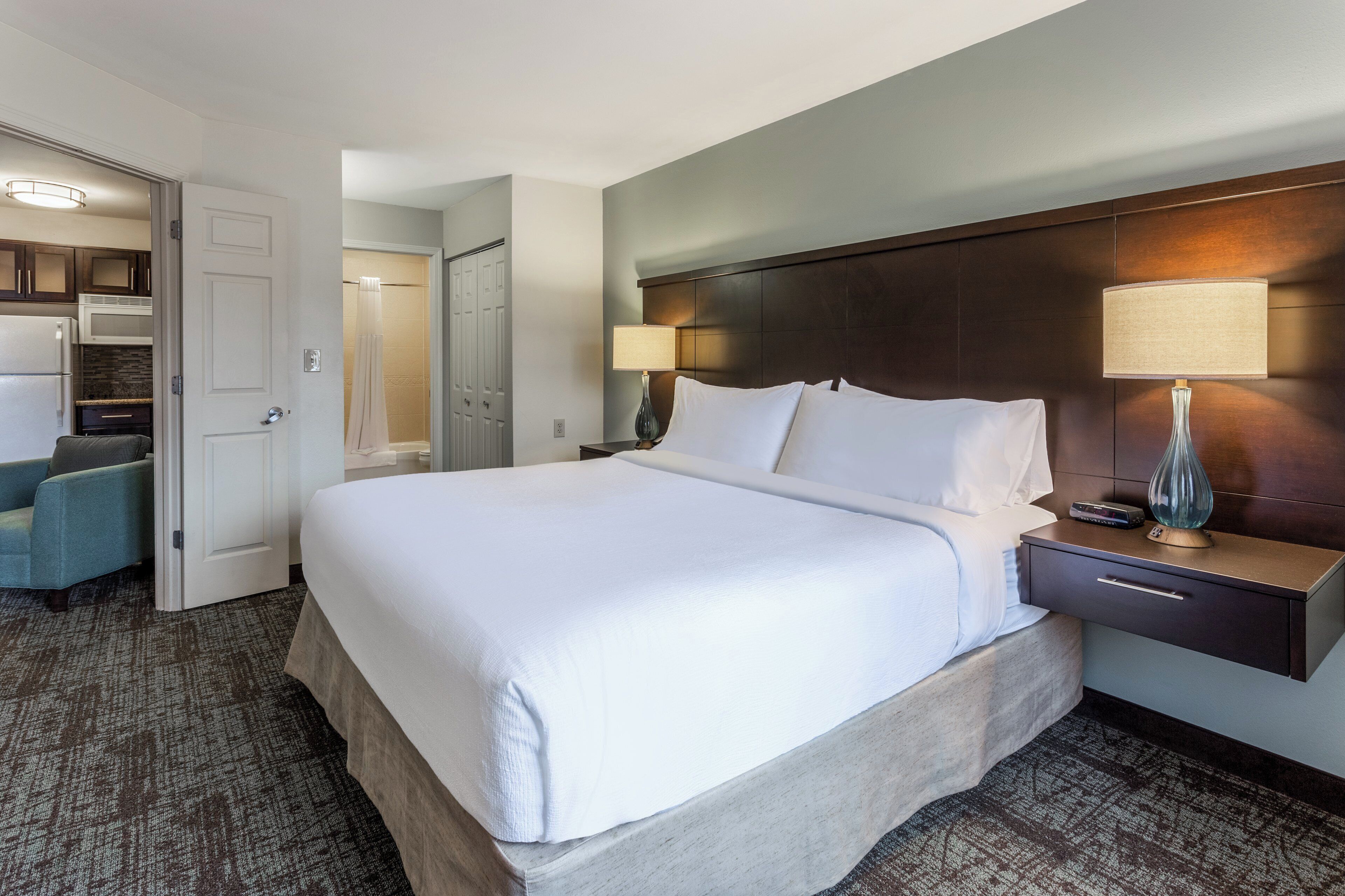 A hotel room with a large bed, a bedside table with a lamp, a nightstand, a wardrobe, and a bathroom with a shower curtain.