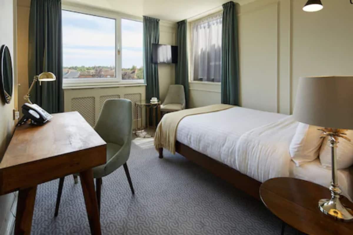 A hotel room with a bed, a desk with a telephone, a chair, a small table, a lamp, and a TV on the wall.