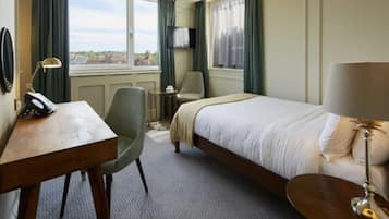 Habitación de hotel con cama, escritorio con teléfono, silla, mesita, lámpara y televisor en la pared.