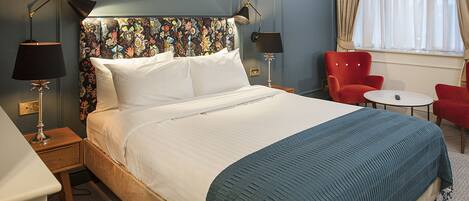 Habitación de hotel con una cama grande, dos lámparas de noche, un sillón rojo, una mesita redonda y un ventanal con cortinas.