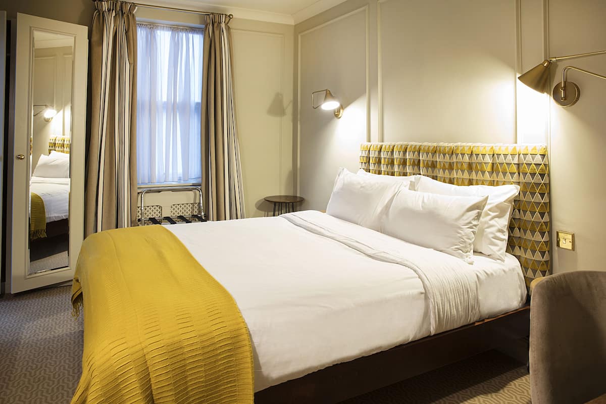 A hotel room with a bed, a mirror, a window with curtains, and a bedside lamp.