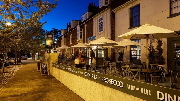 A nighttime outdoor dining area with tables and umbrellas, a signboard with the name 'PROSECCO RUMP & WADE', and a brick wall. - Cromwell Hotel Stevenage (Stevenage)
