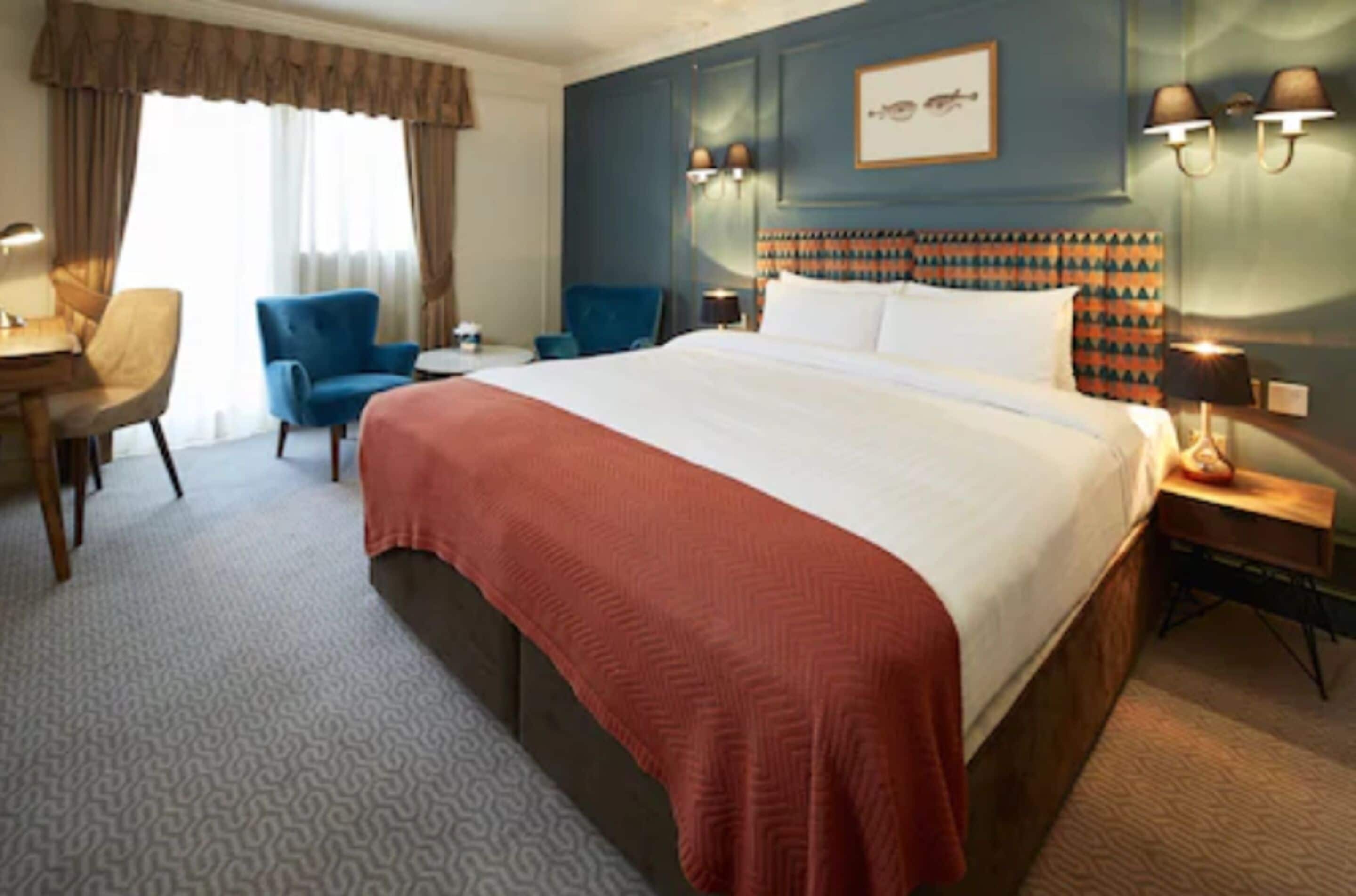 Quarto de hotel com uma cama grande, uma mesa, duas cadeiras e uma mesinha de cabeceira com luminárias.
