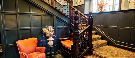 A grand staircase with ornate woodwork, a plush orange armchair, patterned cushions, and a vase of flowers.