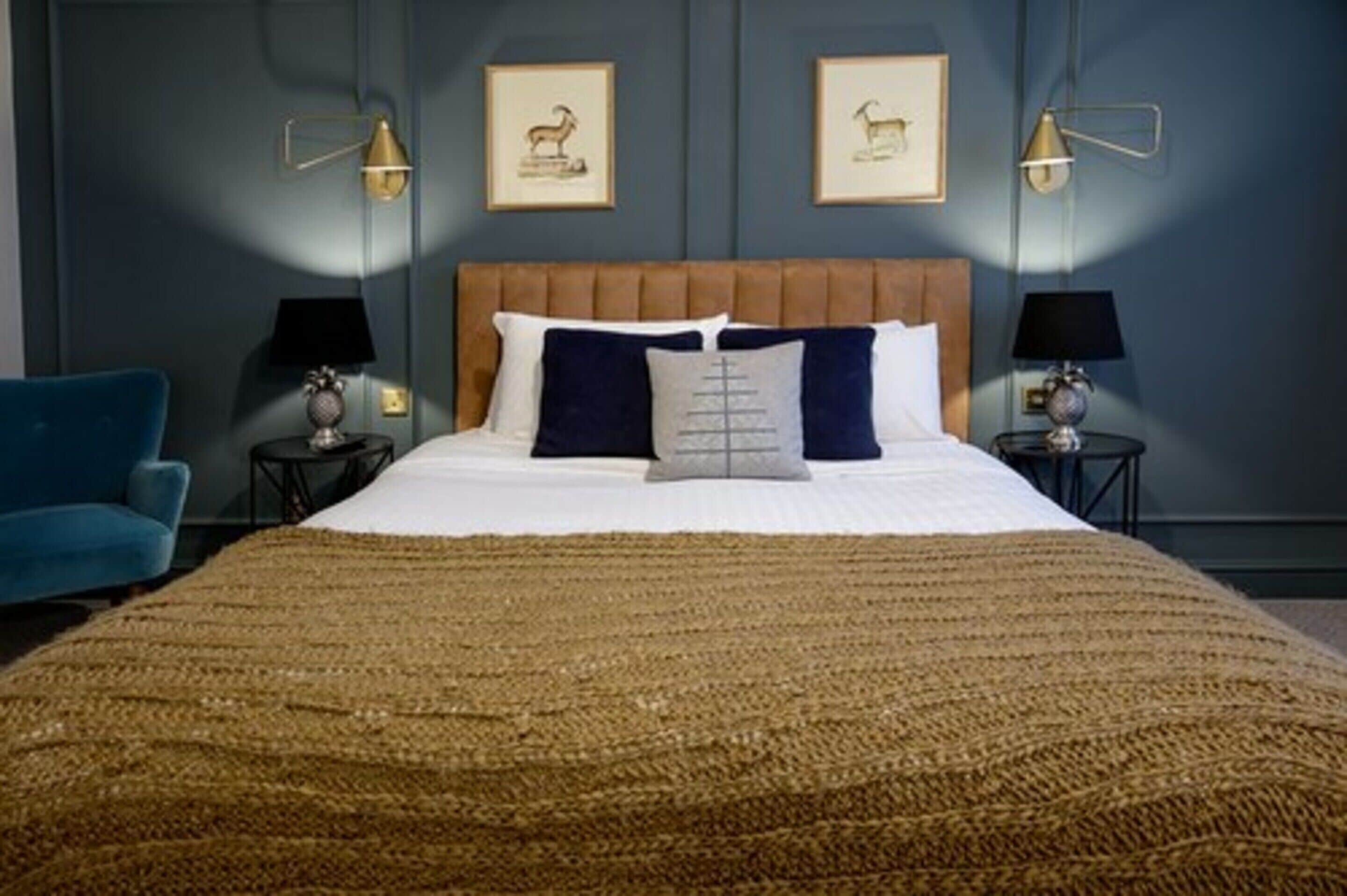 A bedroom with a large bed, two bedside tables with lamps, a blue armchair, and framed pictures on the wall.