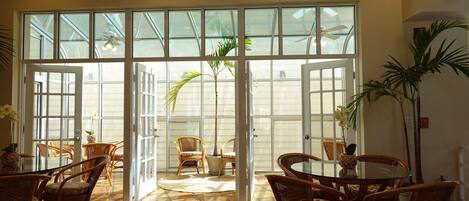 A spacious indoor area with a large glass door leading to an outdoor patio. Inside, there are wicker chairs and a round table, with potted plants and a ceiling fan visible.