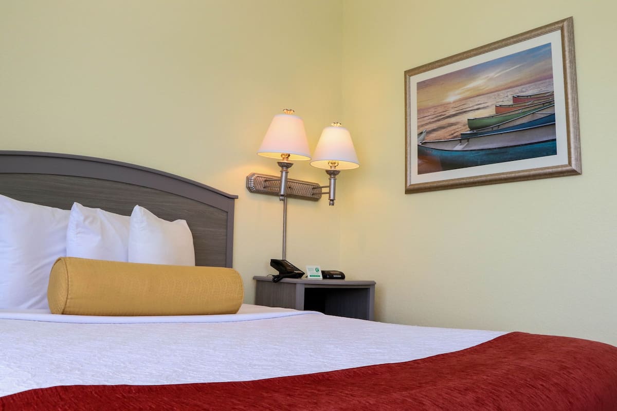 A neatly made bed with white and red linens, a small bedside table with a lamp, a telephone, and a framed picture of boats on the wall.