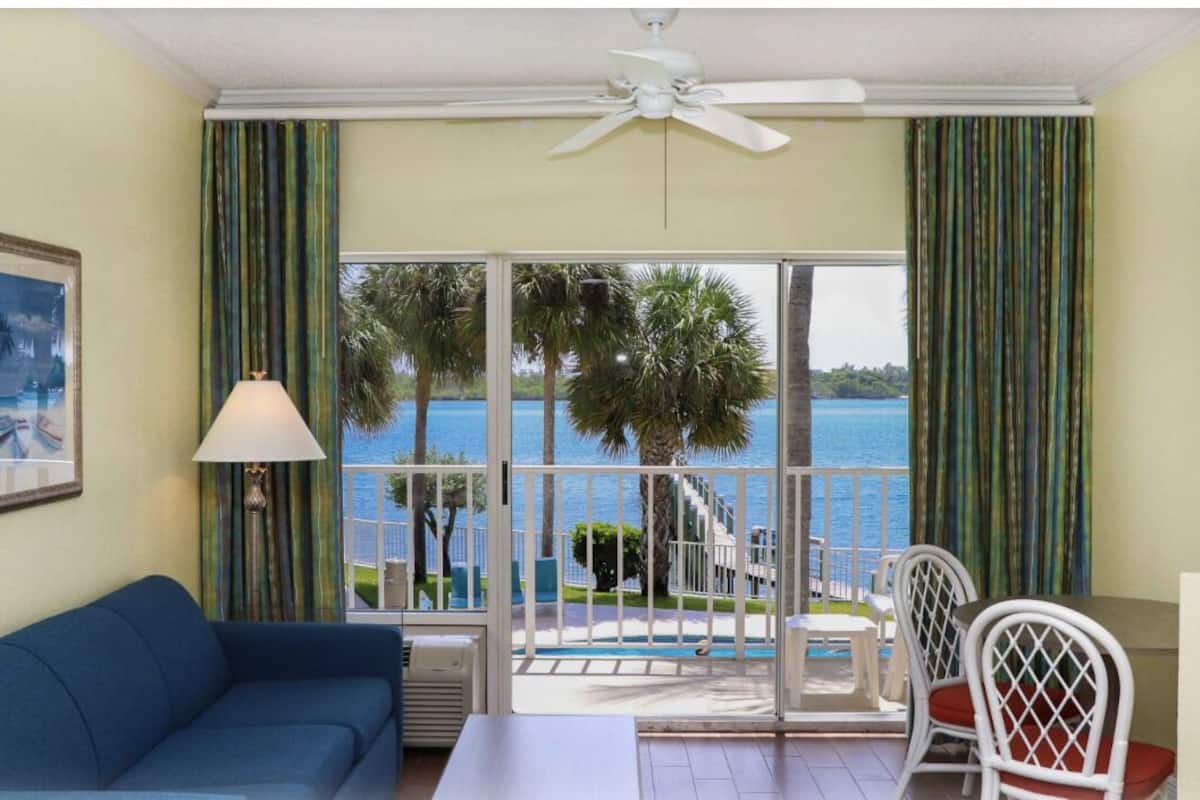 A room with a blue sofa, a table, and chairs, a ceiling fan, and a view of the sea through large windows.