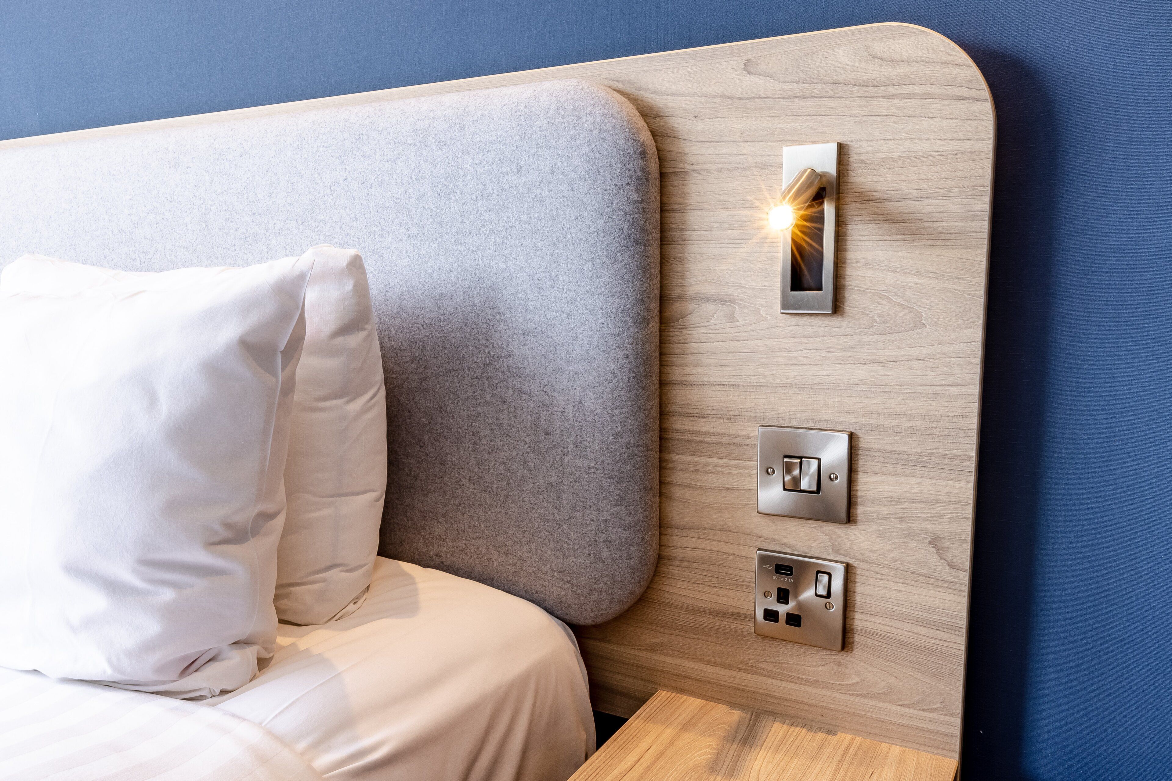 A modern hotel room with a wooden headboard, a bedside table, a white pillow, and a wall-mounted light switch.