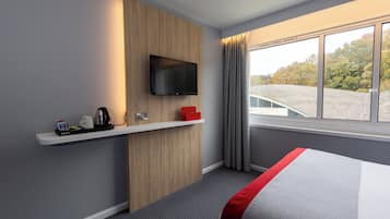 Habitación de hotel con cama, televisión montada, repisa con cafetera y ventana con cortinas.
