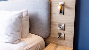 A modern hotel room with a wooden headboard, a bed with white bedding, and a control panel with a light.