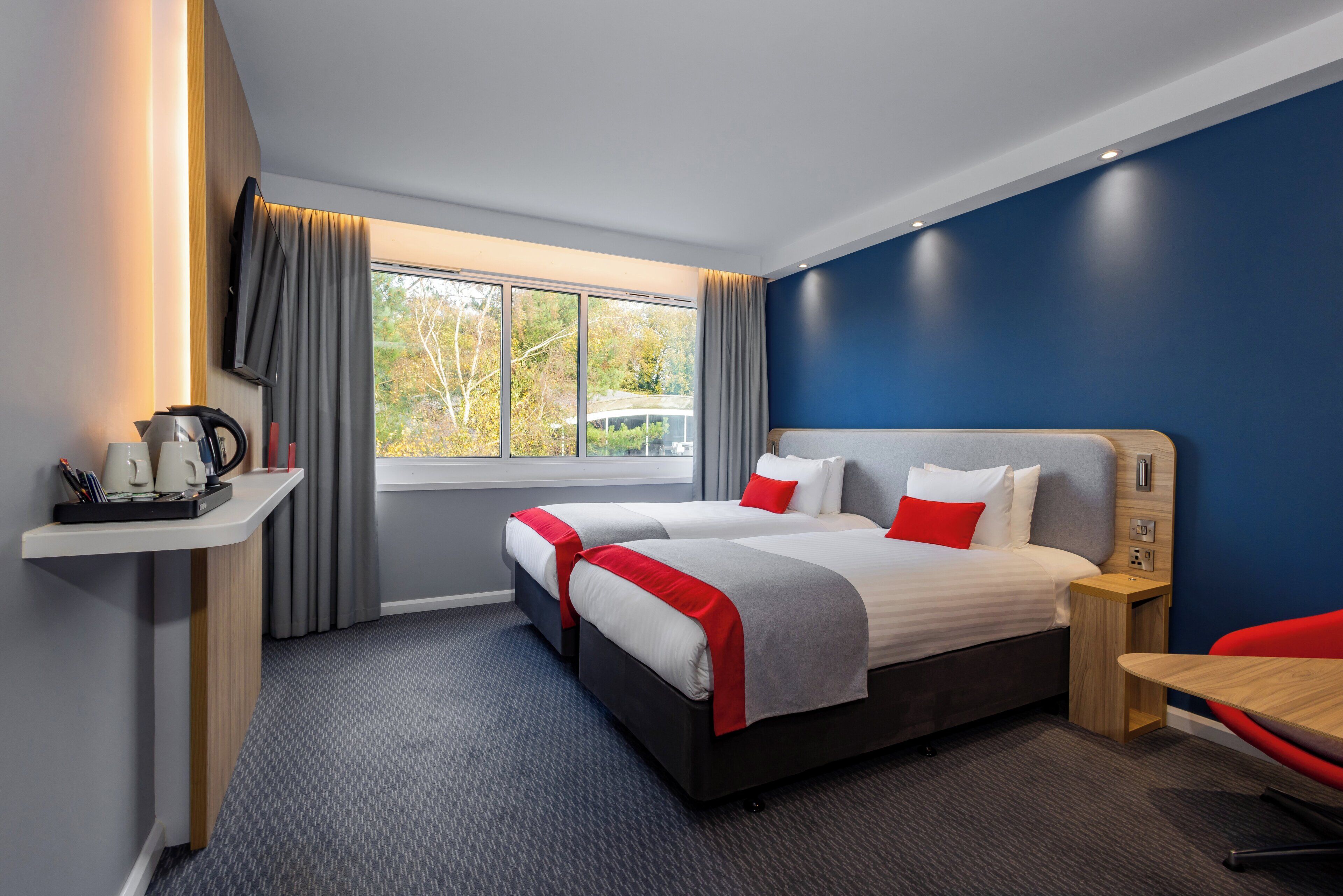A hotel room with two beds, a desk, a chair, a window with curtains, and a coffee maker on a side table.