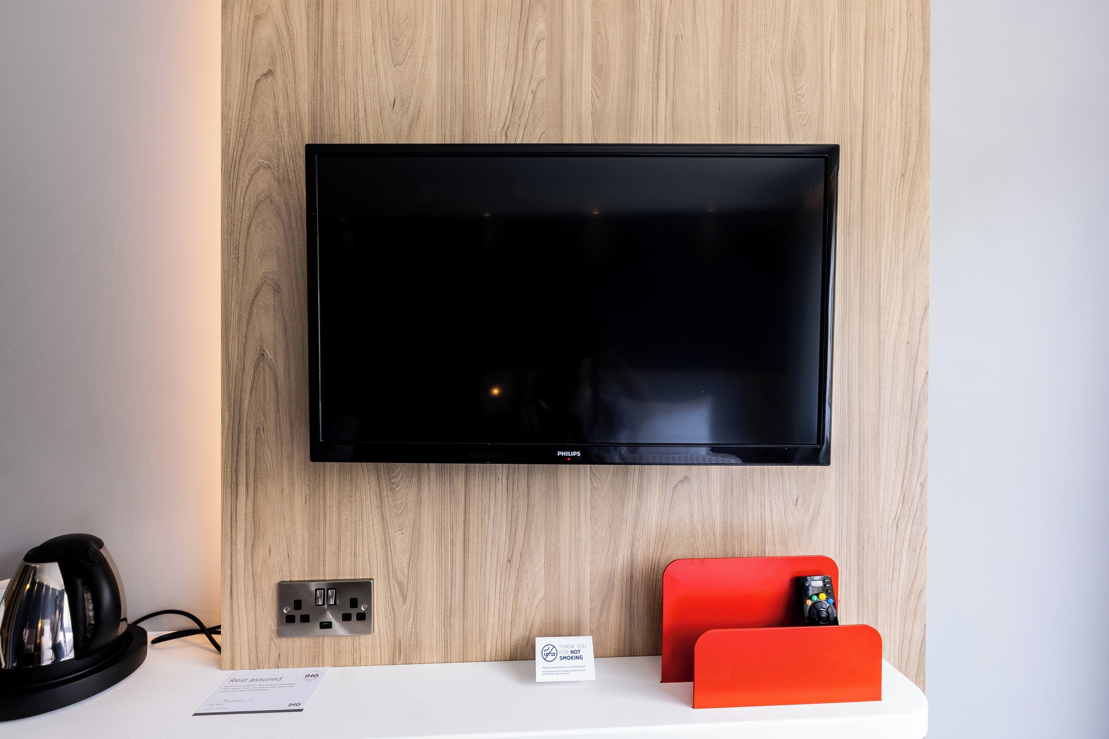 Televisor de pantalla plana montado en una pared de madera, un enchufe, una tetera, una estación de carga roja y un cartel que dice 'Gracias por no fumar'.