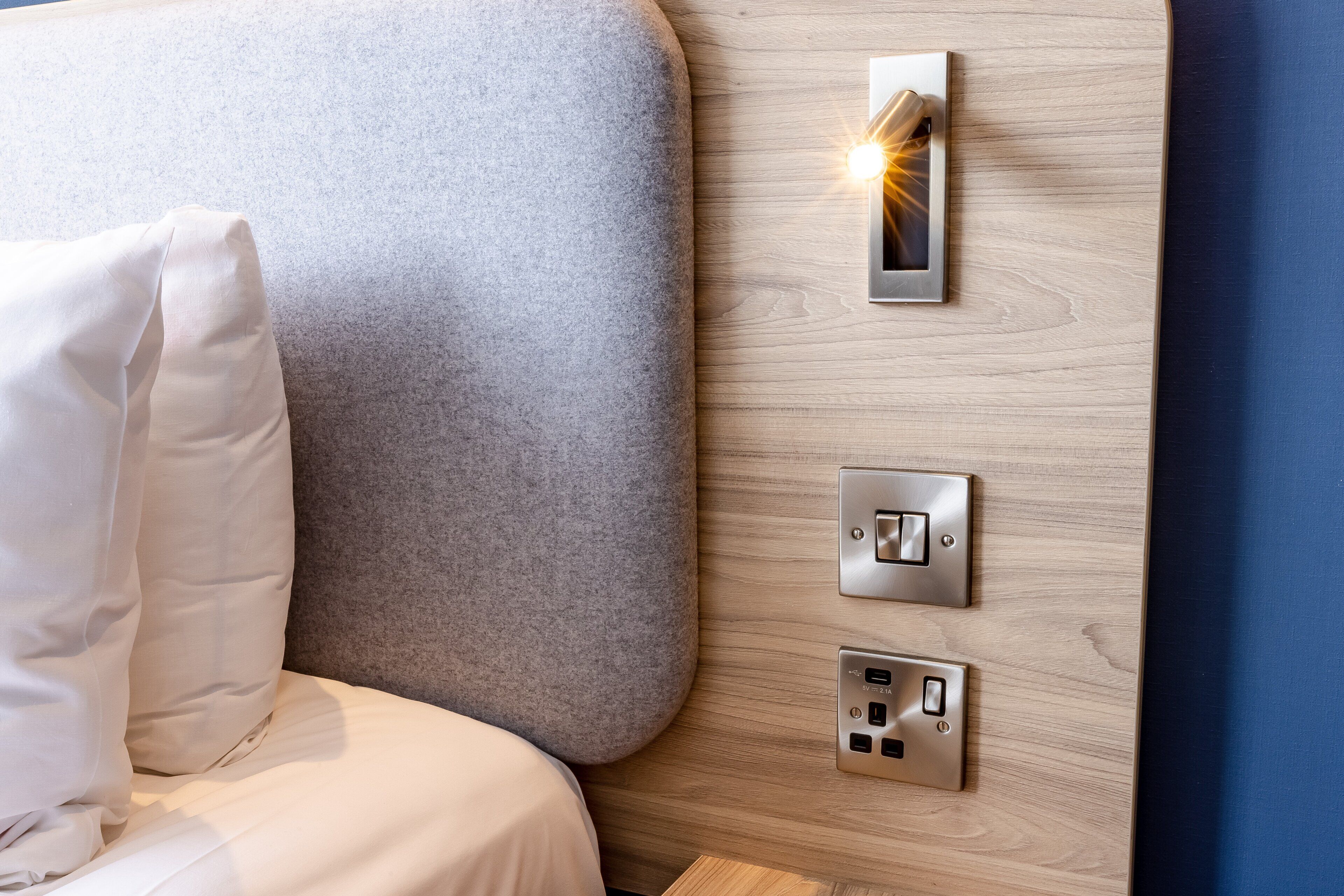 A bed with a grey headboard, a white pillow, and a wall-mounted control panel with a light.