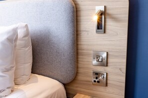 A bed with a grey headboard, a white pillow, and a wall-mounted control panel with a light.