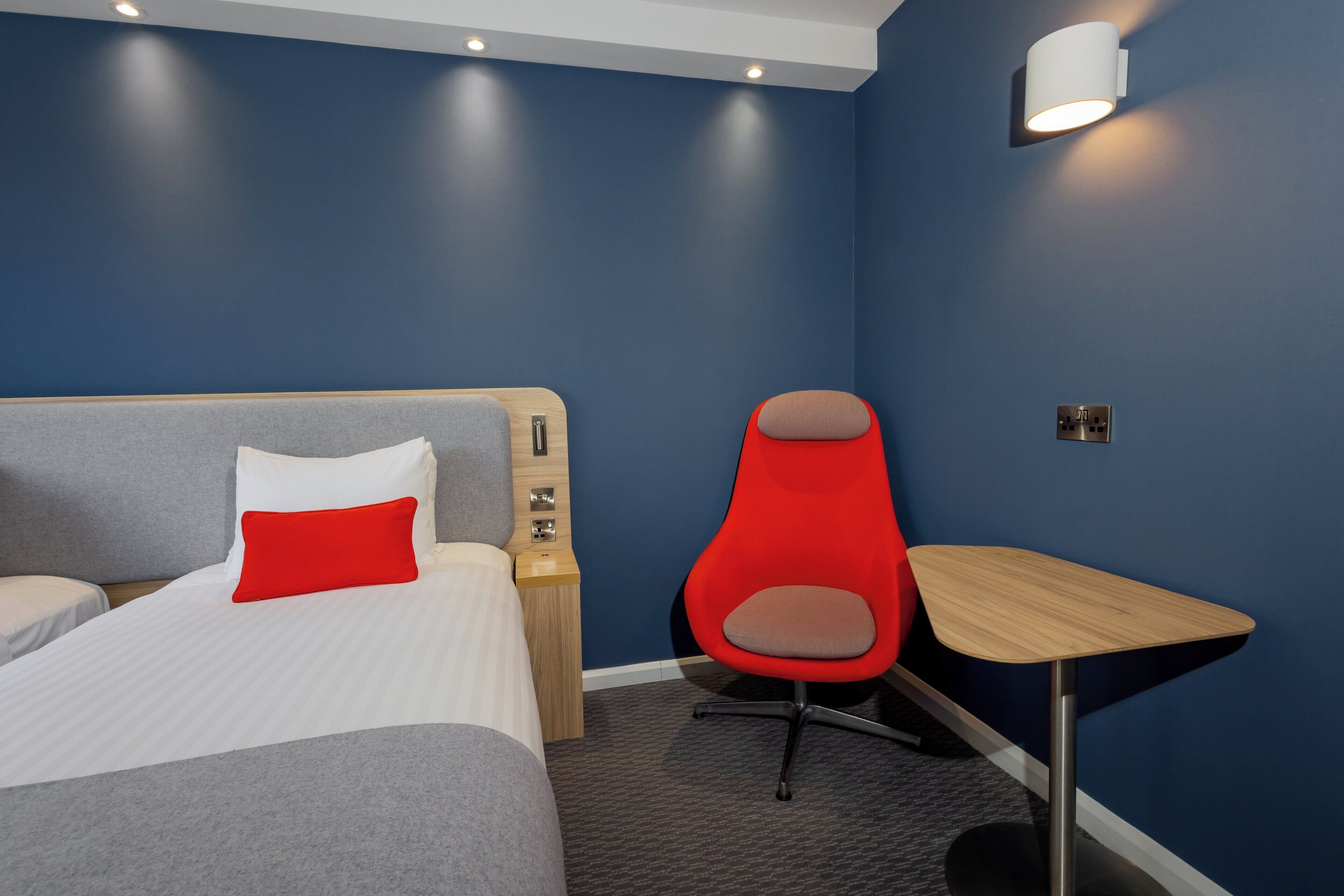 A hotel room with a bed, a red chair, a small wooden table, and a wall-mounted light fixture.