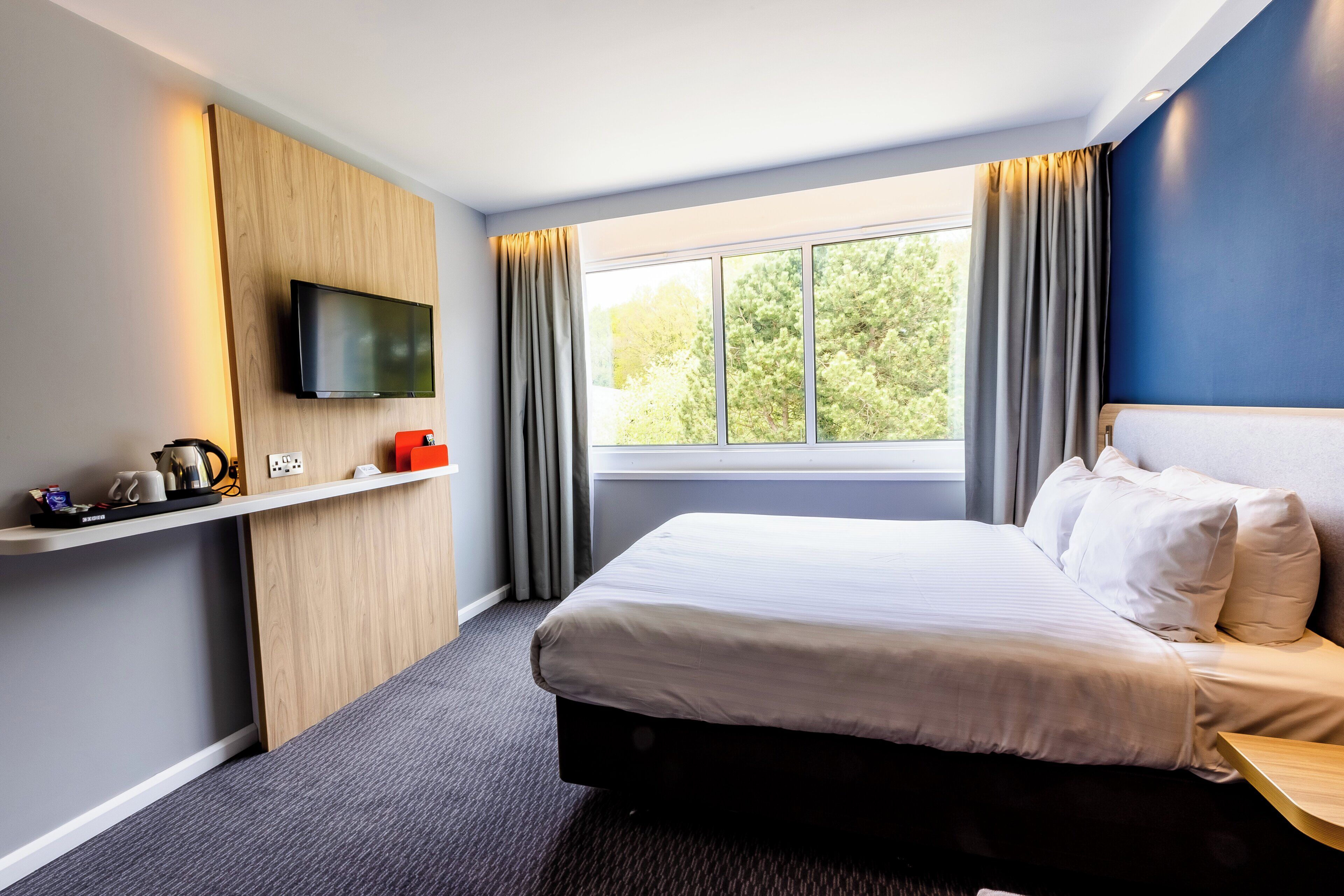A hotel room with a bed, a TV mounted on the wall, a window with curtains, and a small shelf with items.