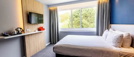 A hotel room with a bed, a TV mounted on the wall, a window with curtains, and a small shelf with items.