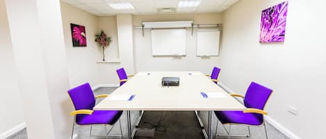 A conference room with a long table, six purple chairs, white walls, and two whiteboards.