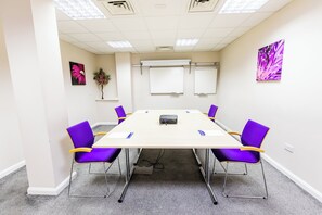 A conference room with a long table, six purple chairs, white walls, and two whiteboards.