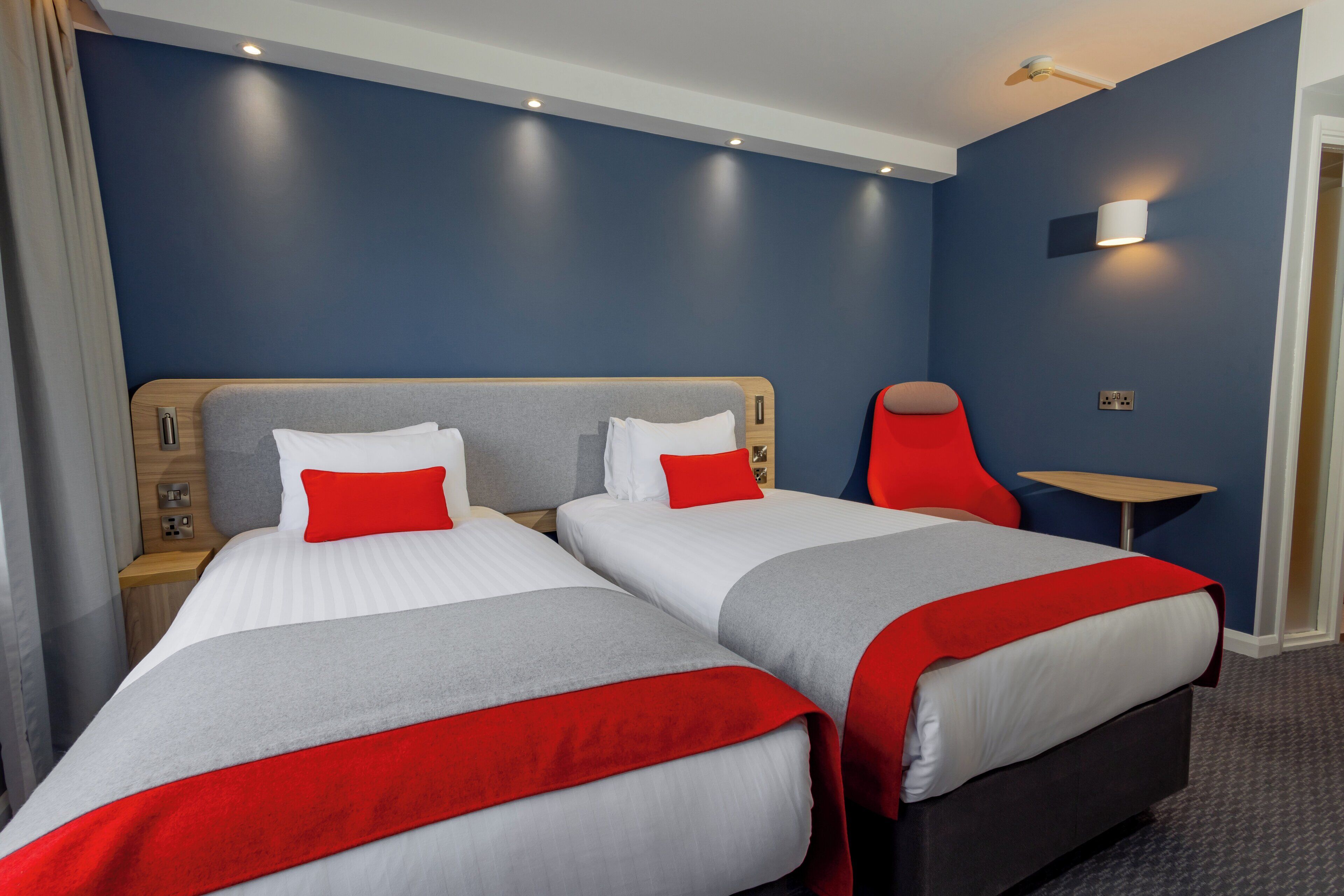 Habitación de hotel con dos camas, cada una con ropa de cama blanca y roja, una silla roja, una mesita pequeña y una pared azul.