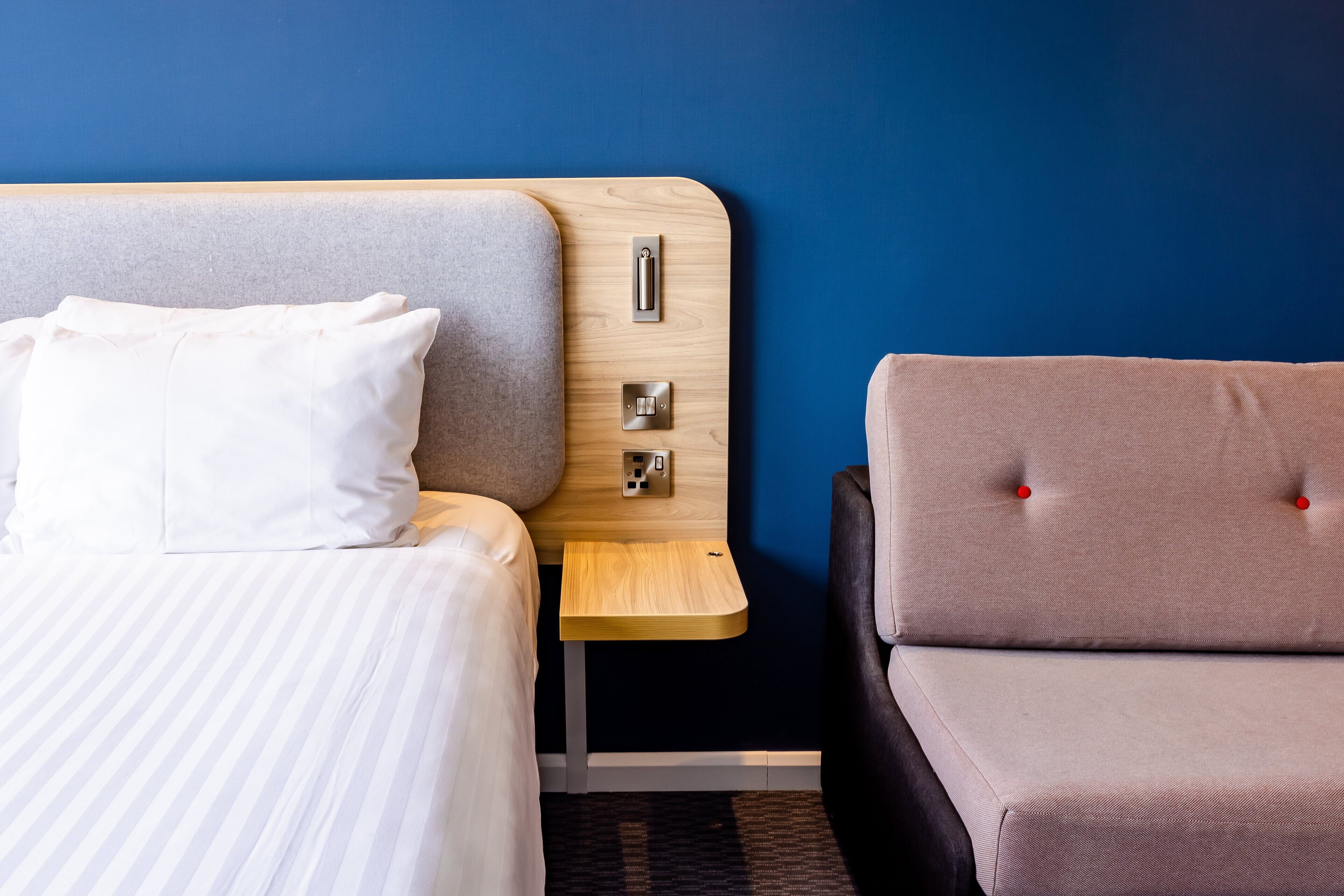 A hotel room with a bed, a bedside table, a chair, and a wall socket.