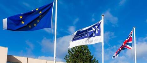 Tres banderas: la bandera de la Unión Europea, la bandera del Reino Unido y una bandera con un logotipo y texto.