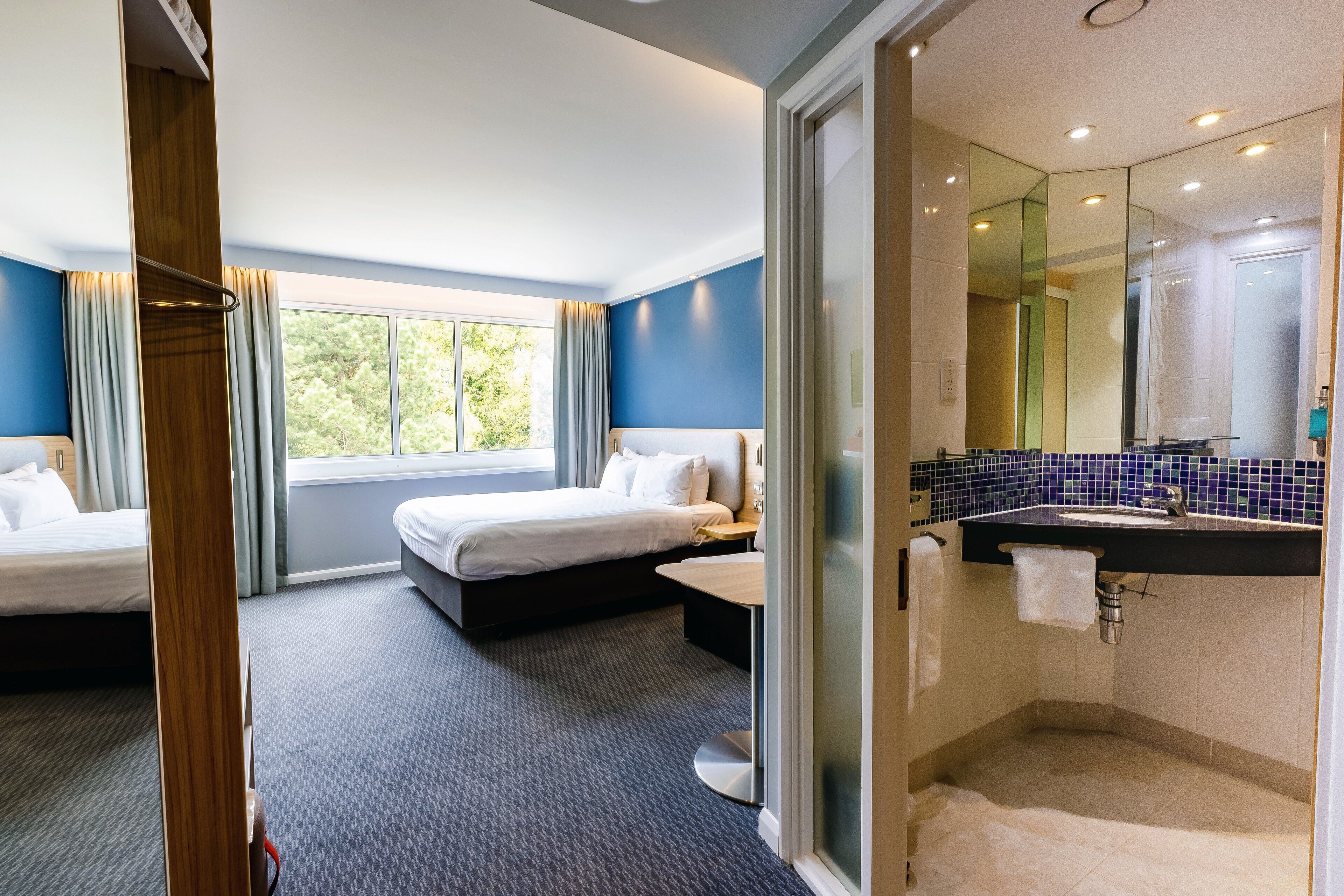 A hotel room with two beds, a bathroom with a mirror, and a view of trees through the window.