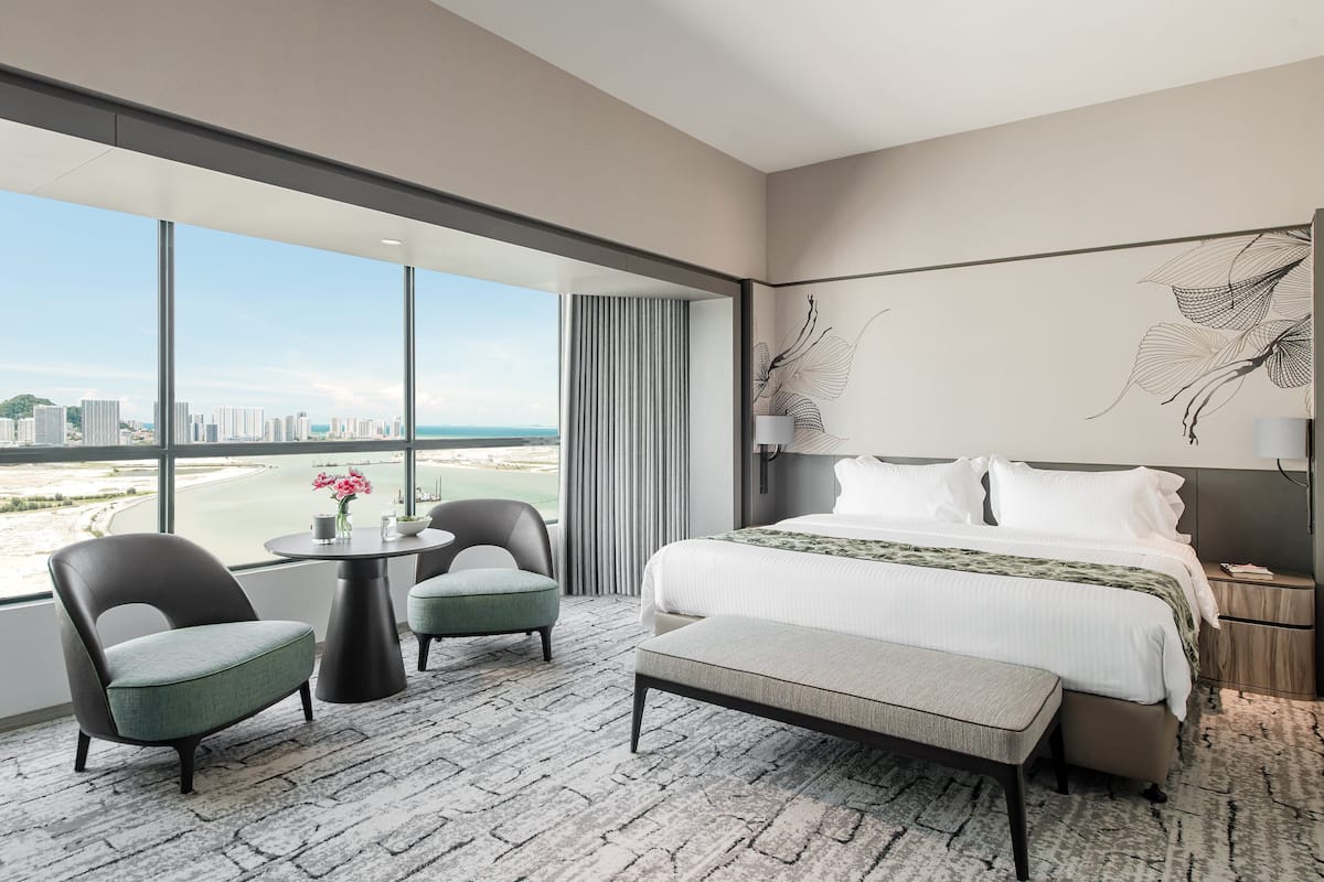 Una habitación de hotel moderna con una cama grande, una zona de estar con dos sillones, una mesita con un jarrón de flores y vistas a la ciudad a través de amplias ventanas.
