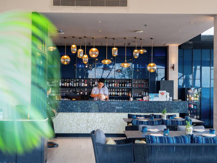 Un espace bar moderne avec un barman, des sièges dotés de coussins bleus et une variété de bouteilles exposées sur des étagères.