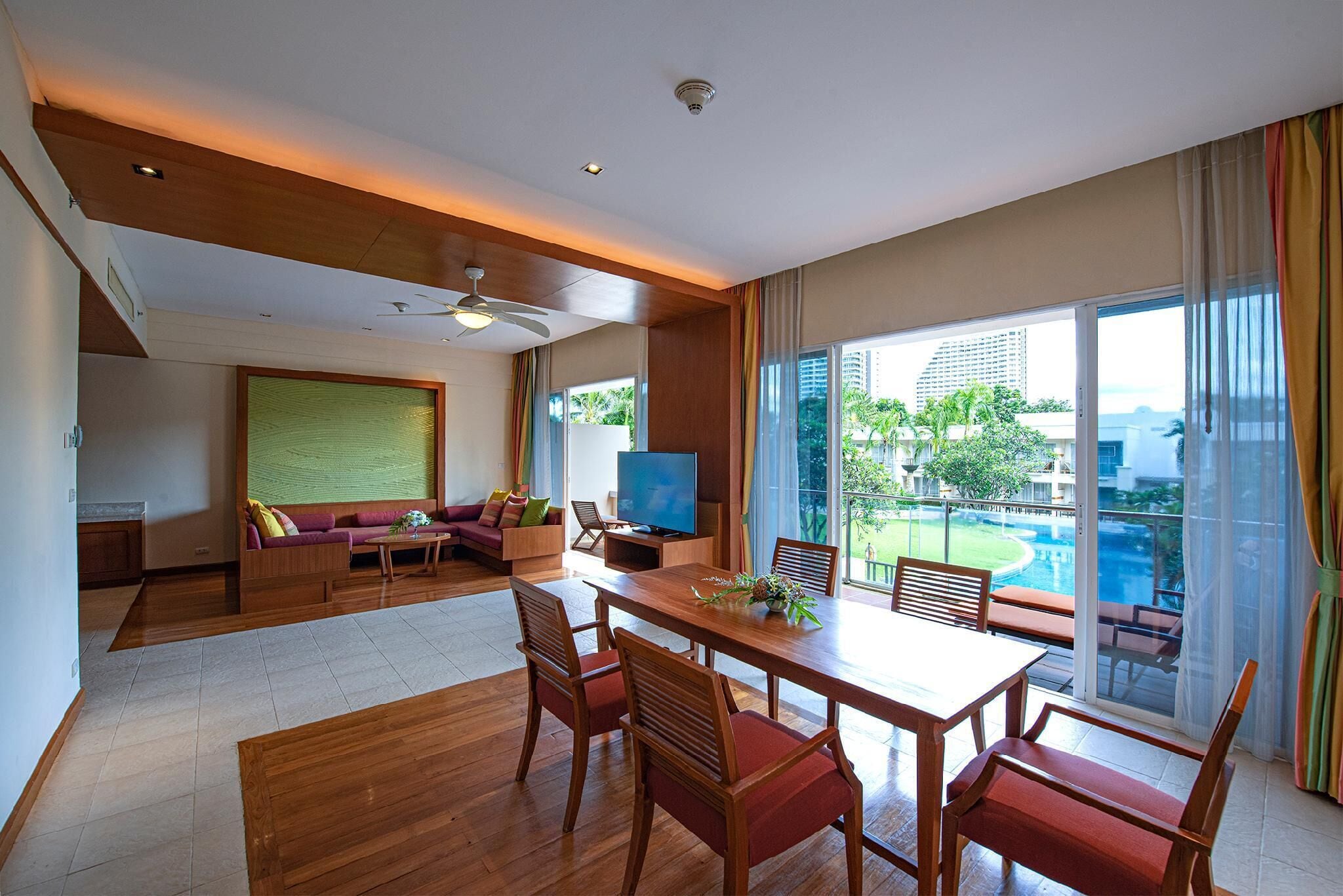 A spacious living area with a wooden dining table, red cushioned chairs, a sofa, and a television. Large windows offer a view of a pool and greenery.