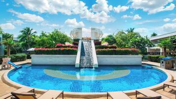 Relax by the serene pool surrounded by lush greenery and palm trees, complete with a central waterfall feature and lounge chairs.