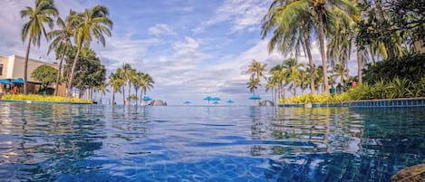 A clear swimming pool with palm trees and umbrellas around it.