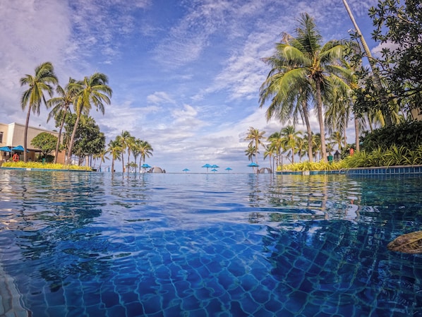 A clear swimming pool with palm trees and umbrellas around it.