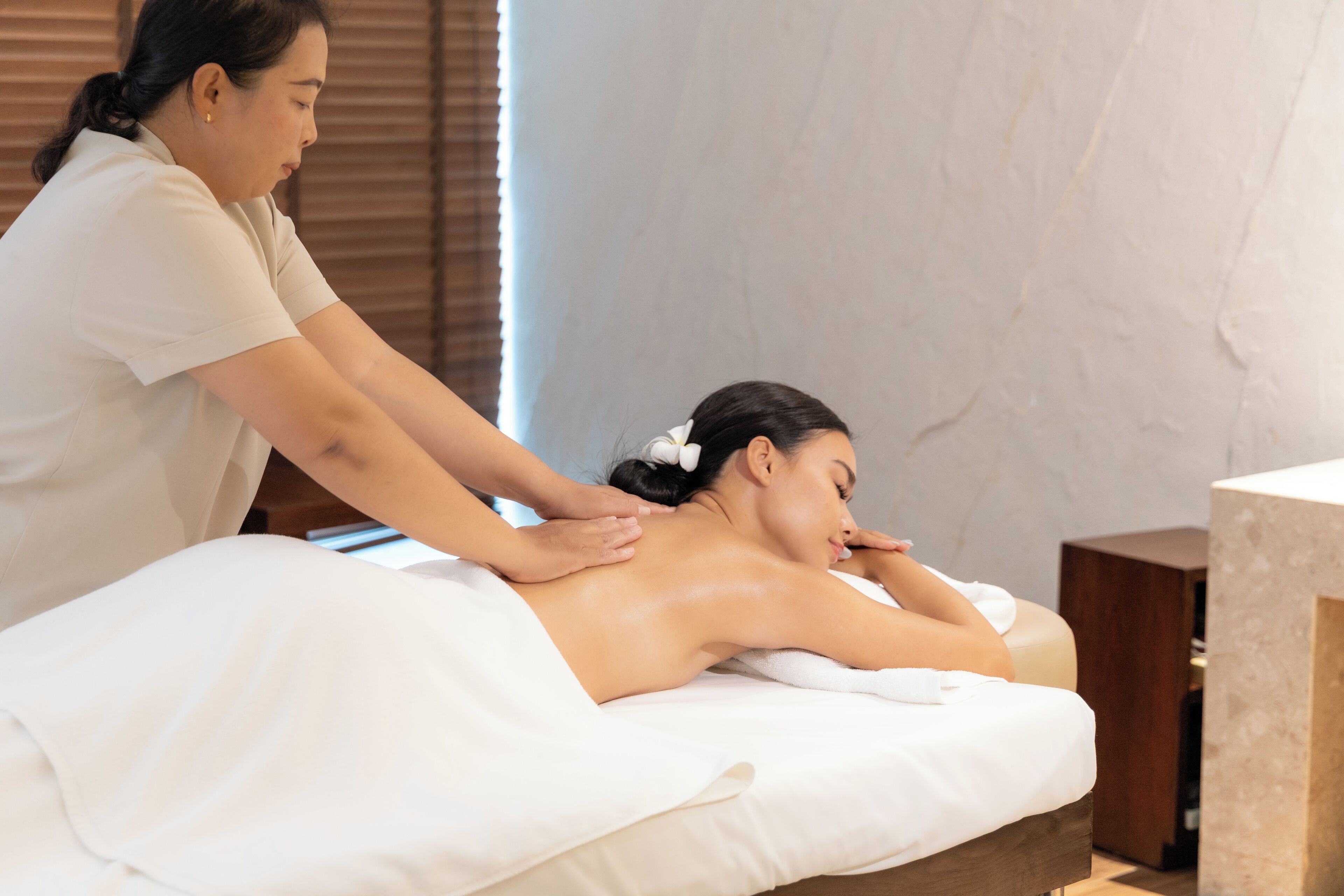 Eine Massageanwendung in einer Wellnessumgebung mit Massagetisch, Holzbeistelltischen und einer weißen Wand.