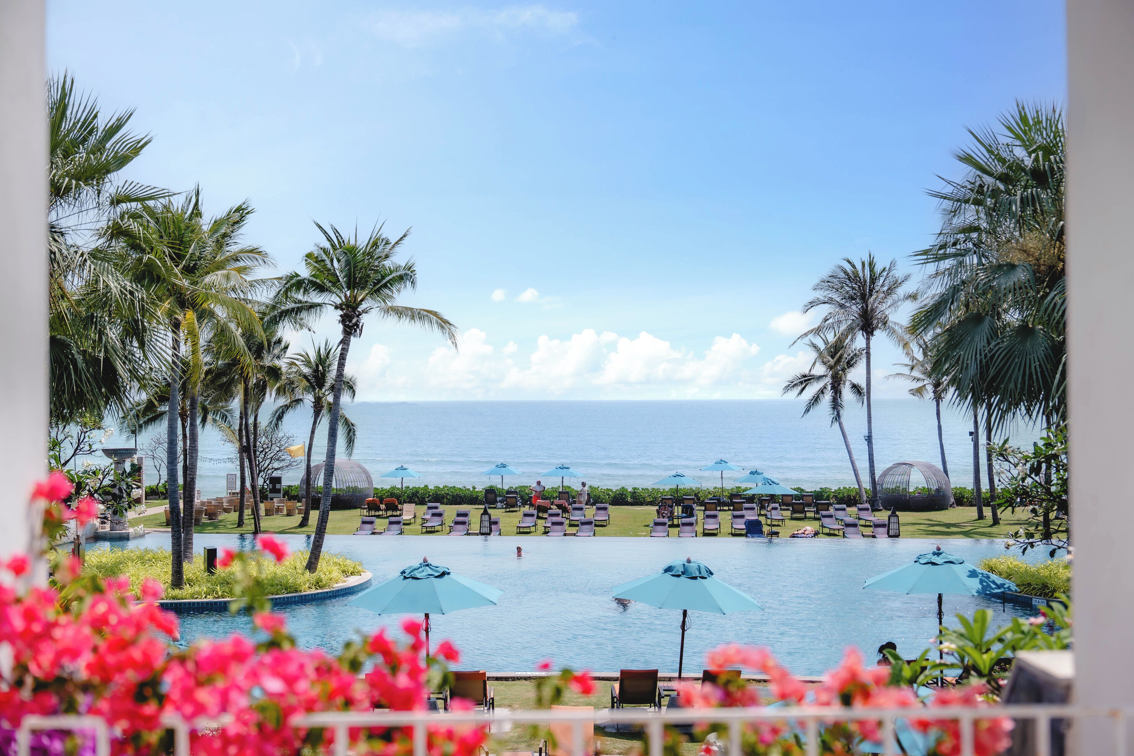 A resort pool with lounge chairs and umbrellas, surrounded by palm trees and a view of the ocean.