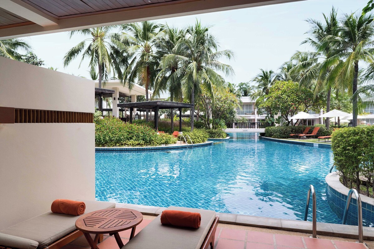 A pool area with lounge chairs and a covered seating area, surrounded by palm trees and a clear blue pool.