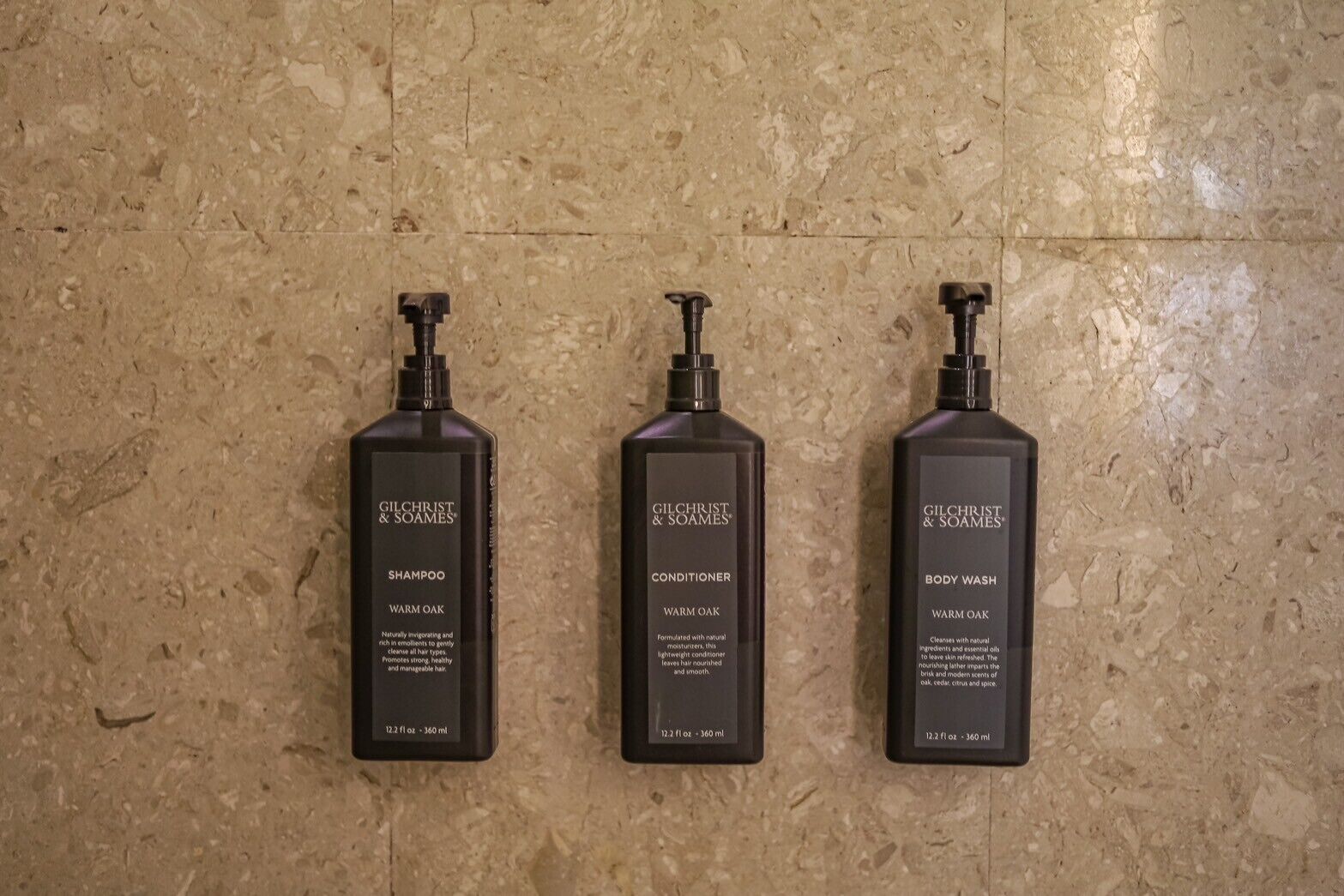 Three Gilchrist & Soames products: shampoo, conditioner, and body wash, all in black bottles with pump dispensers, against a textured beige wall.