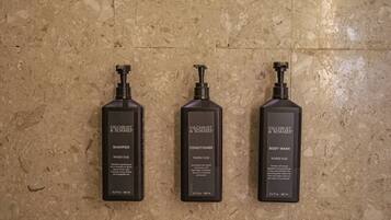 Three Gilchrist & Soames products: shampoo, conditioner, and body wash, all in black bottles with pump dispensers, against a textured beige wall.