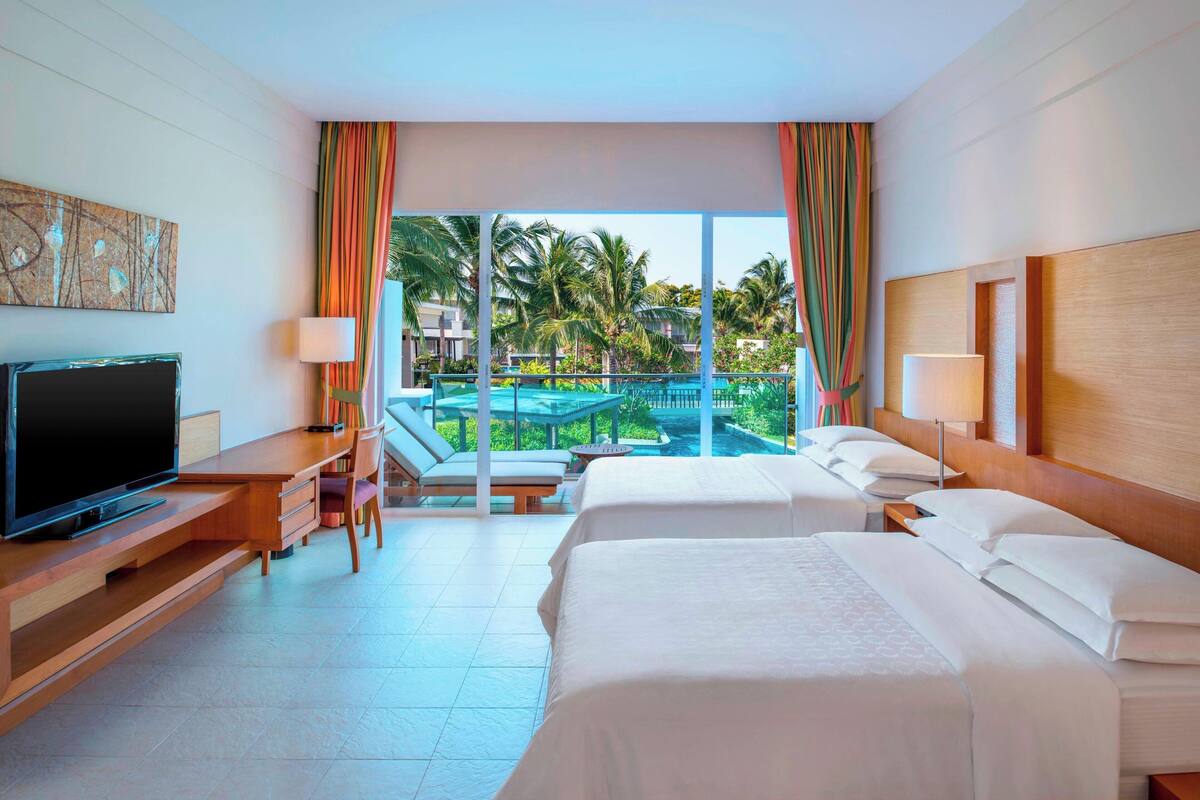 Une chambre d’hôtel avec deux lits, une télévision, un bureau et un balcon donnant sur une piscine et des palmiers.