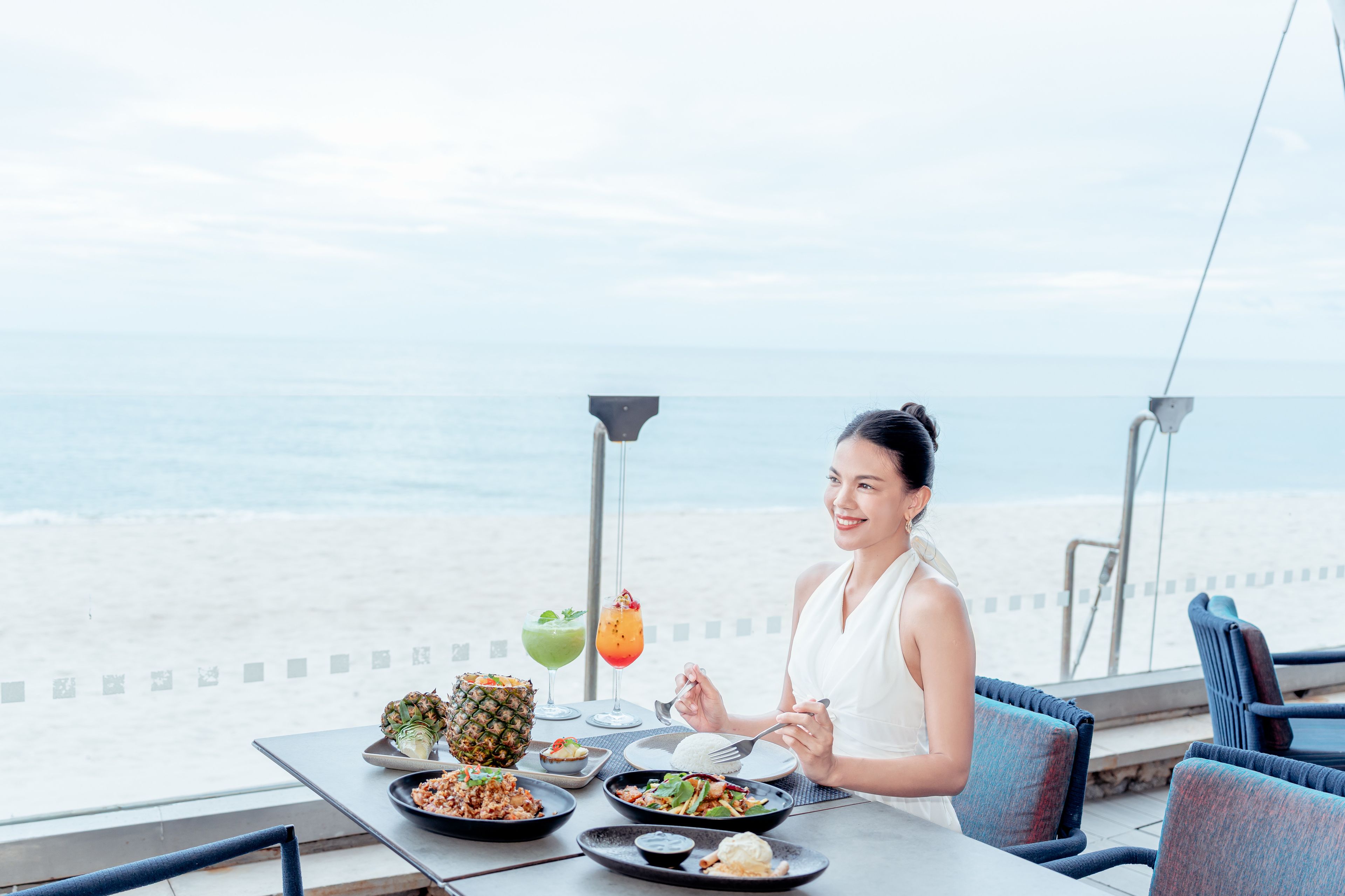 A dining table with various dishes and drinks, overlooking a beach.