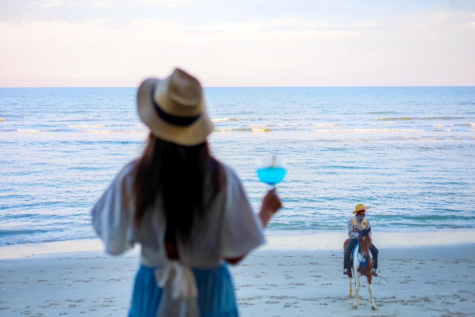 A beach scene with a person in the foreground holding a blue object and another person on horseback in the background.