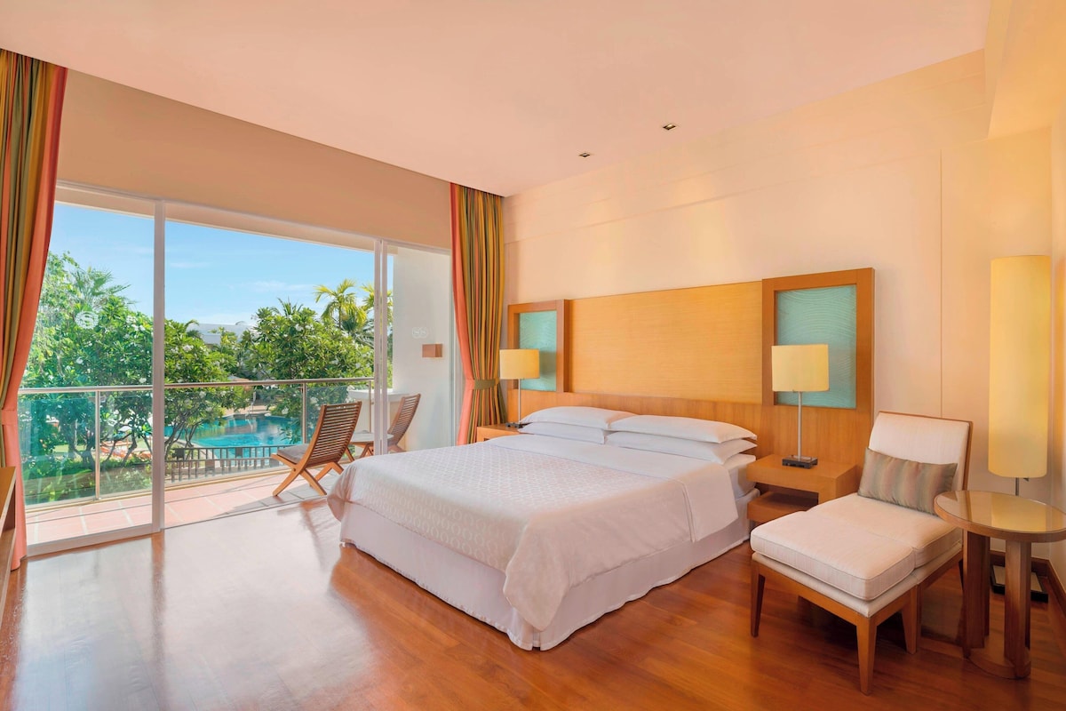 Ein Hotelzimmer mit einem großen Bett, einem Nachttisch, einem Sessel und Blick auf einen Pool.