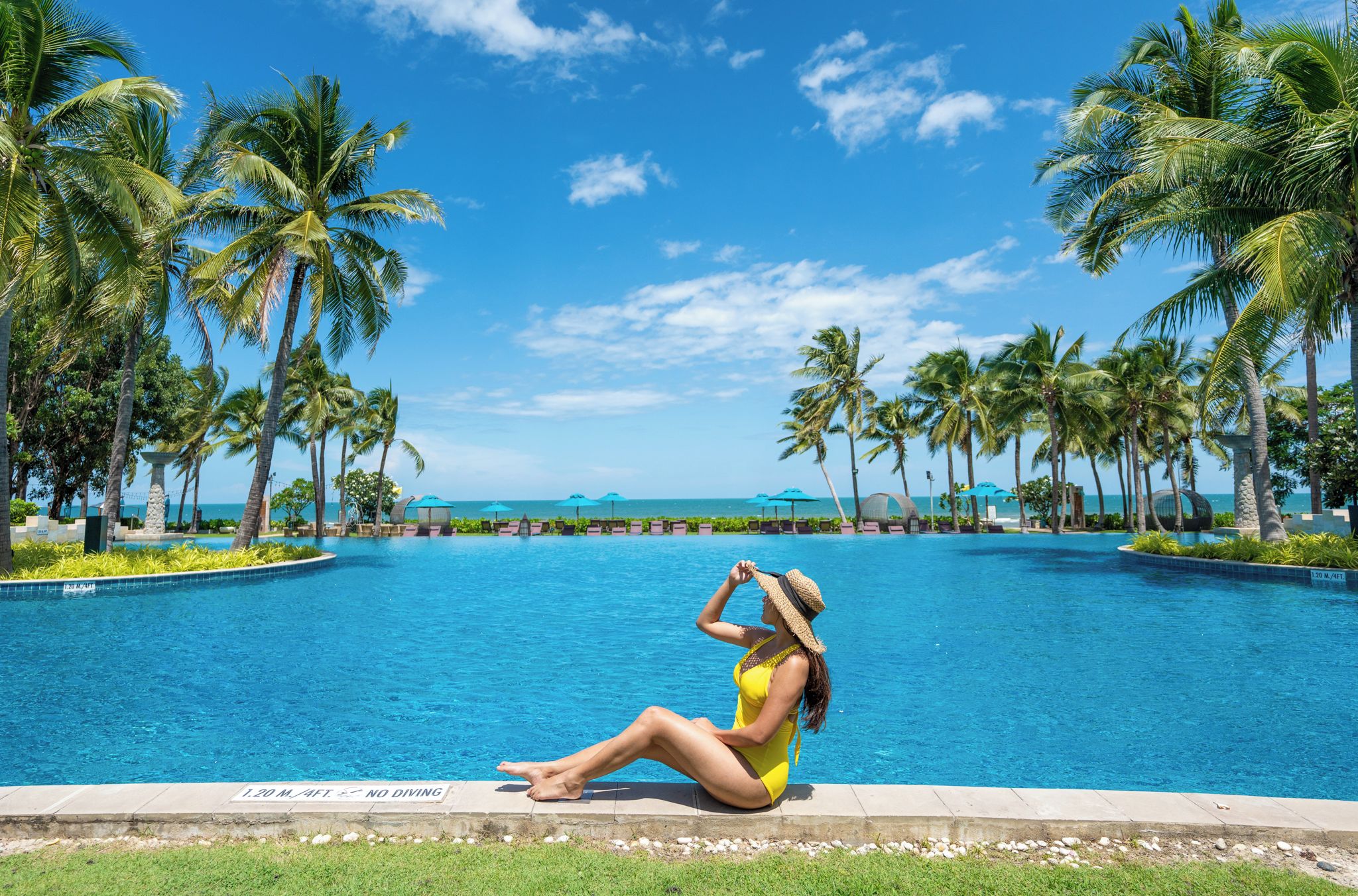 A woman sitting by a pool with palm trees and a clear sky.