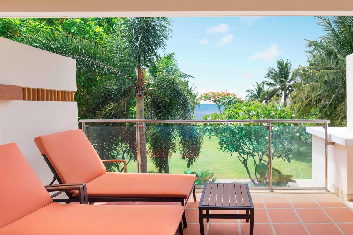 A balcony with two orange chairs and a small table, overlooking a lush garden and a clear sky.