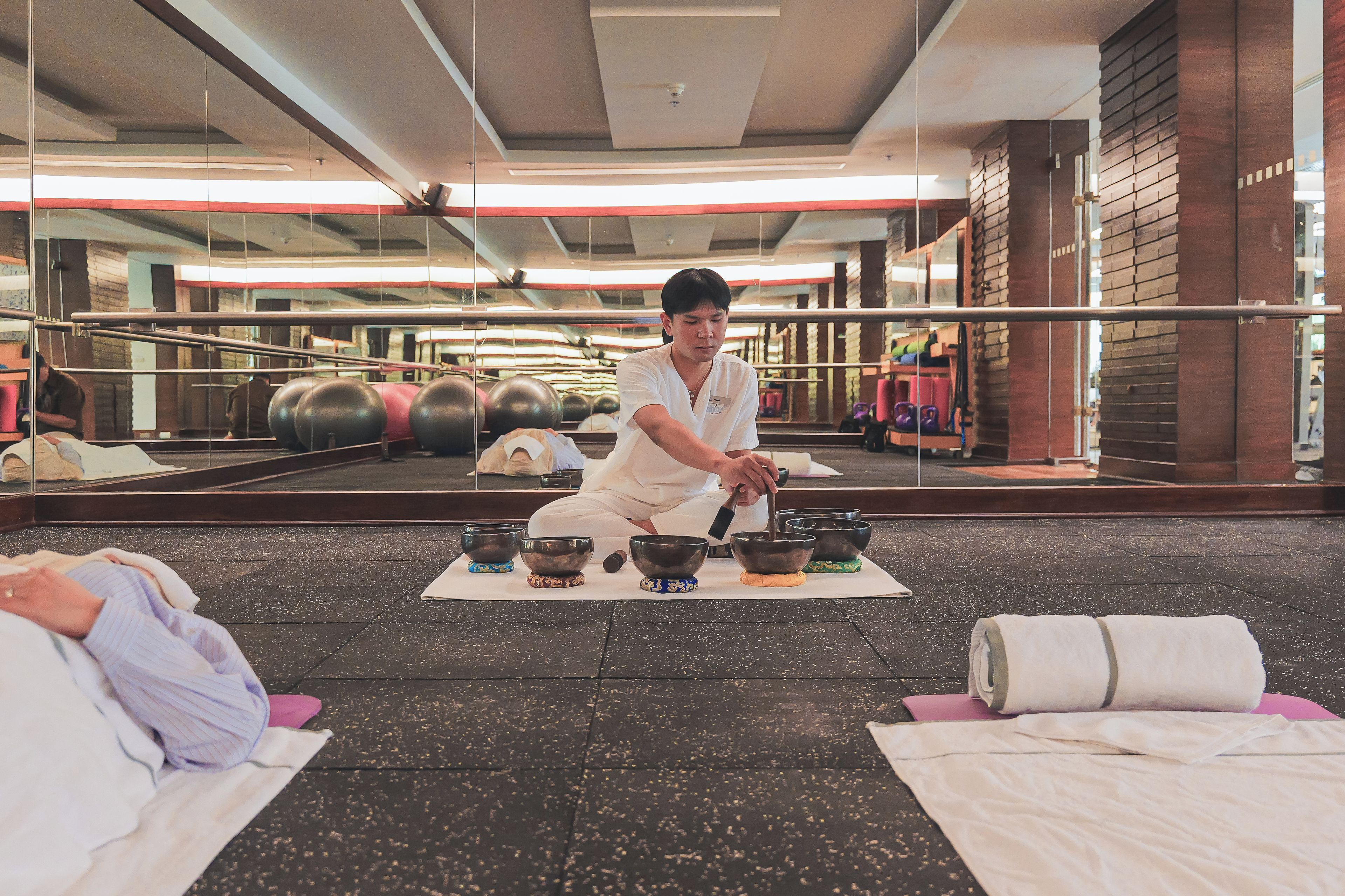 Une personne médite, assise sur un tapis dans une pièce équipée de matériel de sport et dotée d’un grand miroir, et a devant elle un ensemble de bols.