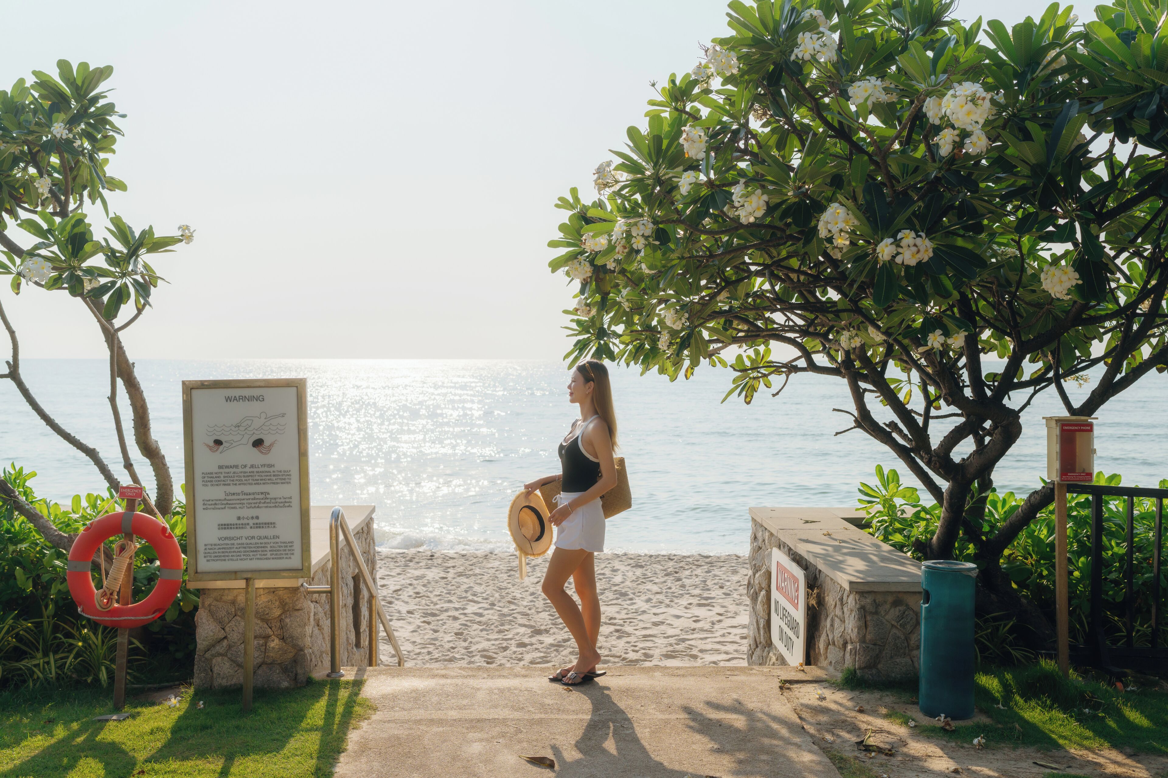 A beachside walkway with a lifebuoy, a warning sign, and a tree with white flowers.