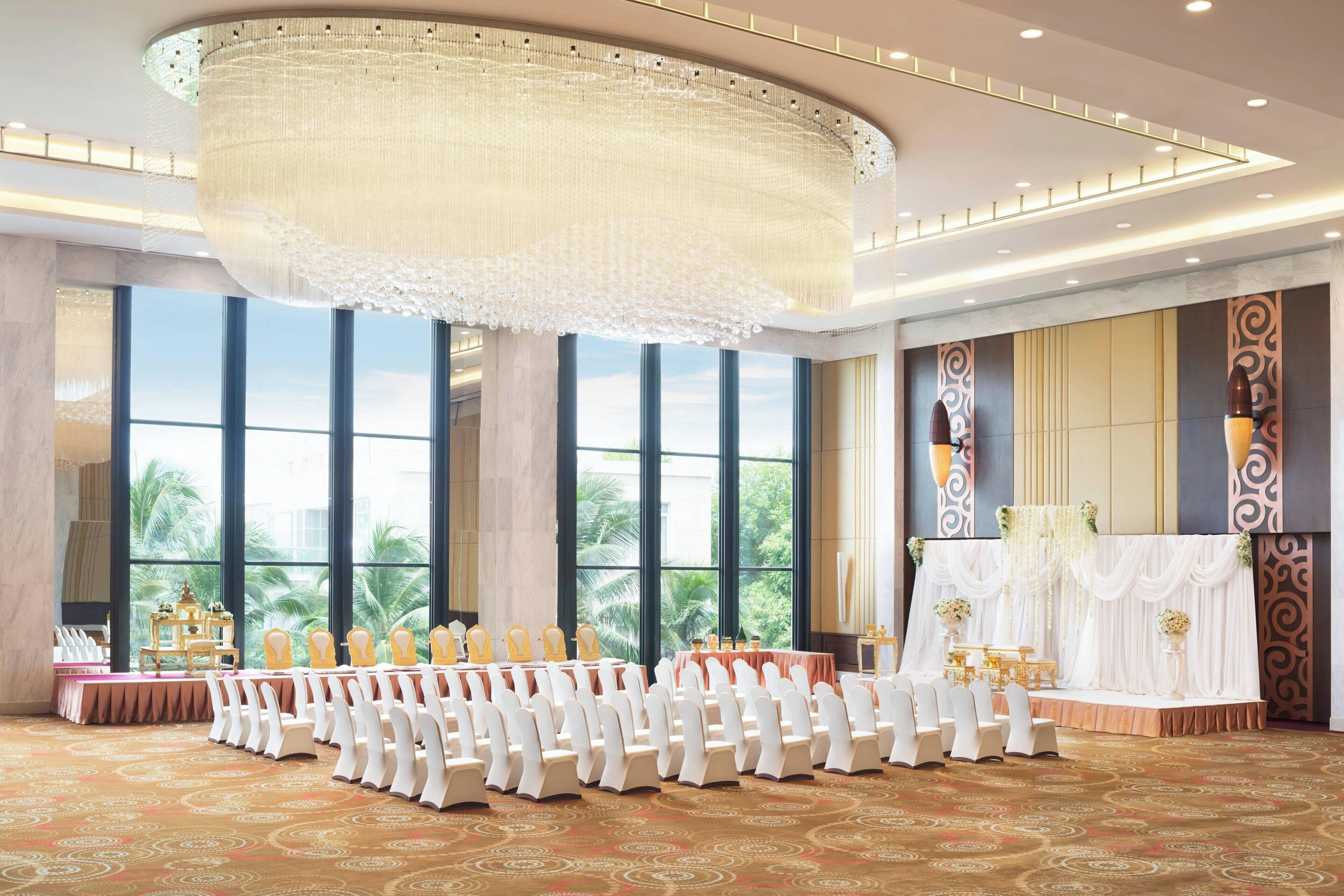 A banquet hall with a large chandelier, long tables with white chairs, and large windows with a view of palm trees.