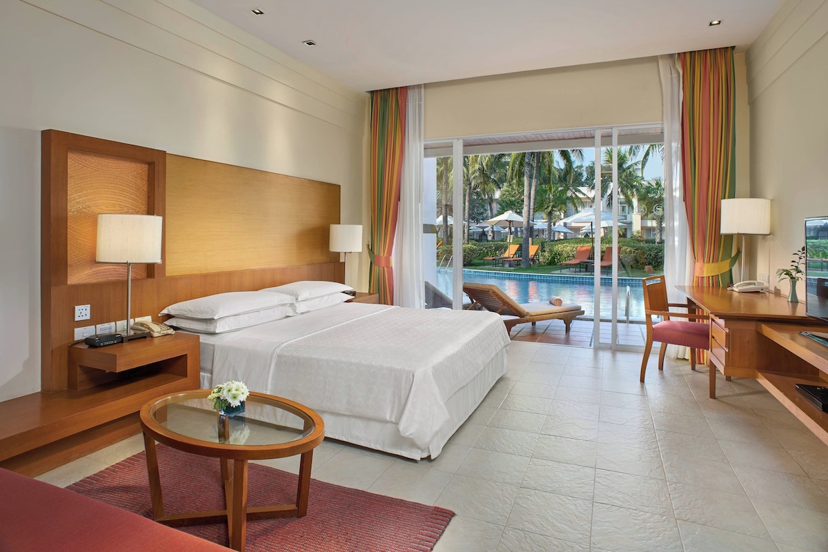 A hotel room with a bed, bedside tables, a desk, a chair, a small table with a vase, and a view of a pool and palm trees outside.